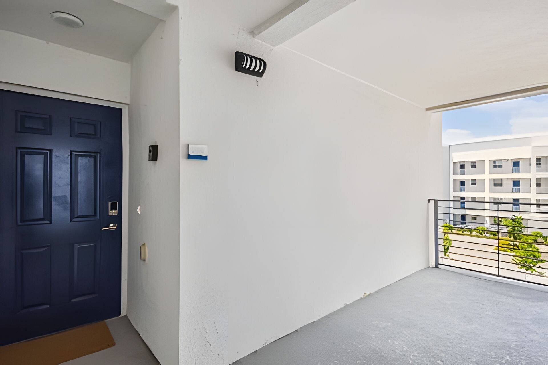 A clean, modern apartment with a dark blue door, a small balcony with a metal railing, and a view of a building in the background.
