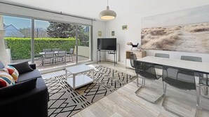 Una sala de estar moderna con un amplio ventanal corredera que da a un patio, un área de comedor con mesa y sillas de vidrio, y un televisor de pantalla plana montado en la pared.