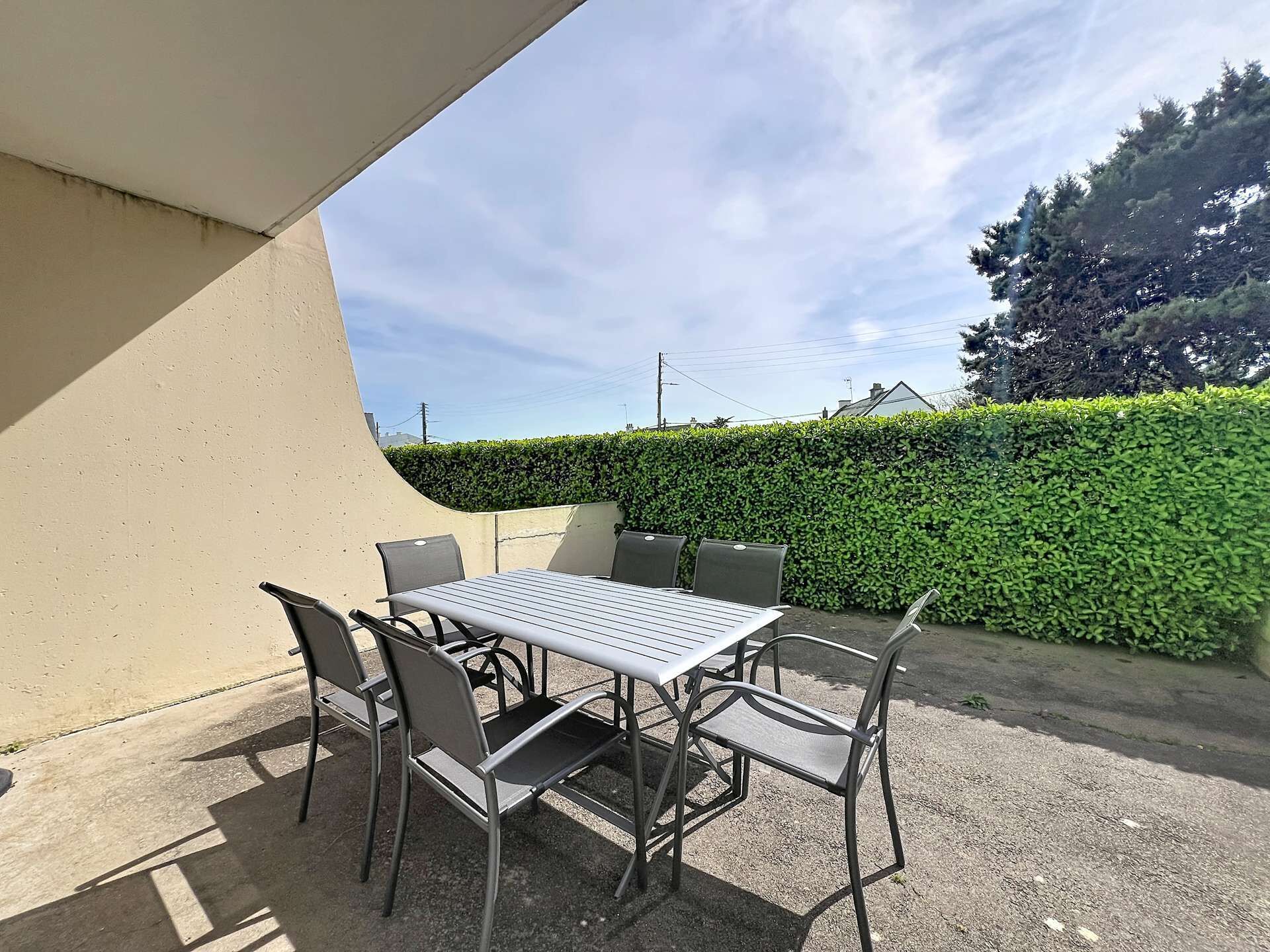 An outdoor patio with a table and chairs, a beige wall, and a hedge in the background.