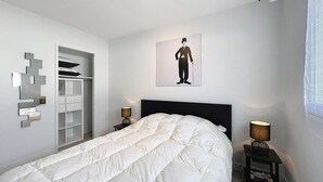Un dormitorio con cama, armario, mesita de noche, lámpara y una obra de arte en la pared.