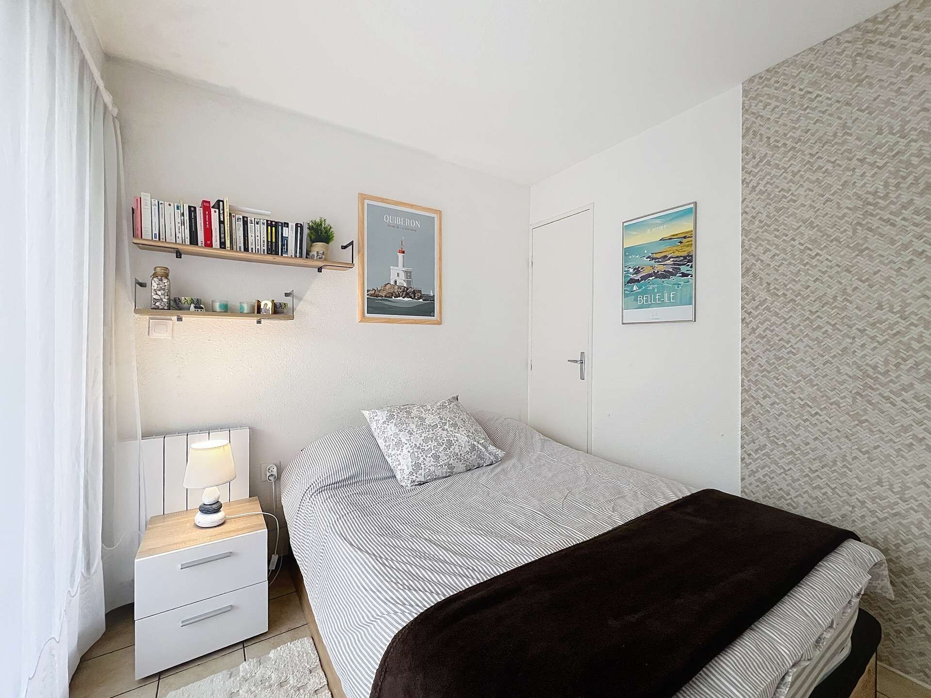 A bedroom with a bed, bedside table, lamp, bookshelf, and two framed pictures on the wall.