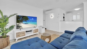 A living room with a large flat-screen TV, blue sofas, a wicker side table, and a potted plant. - 3 Bedroom Apartment Ocean View - Seamist (Kings Beach)