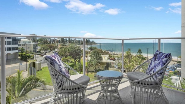 A balcony with wicker chairs and a small table, overlooking a coastal area with buildings and a clear sky. - 3 Bedroom Apartment Ocean View - Seamist (Kings Beach)