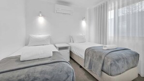 A modern hotel room with two single beds, a small bedside table, an air conditioning unit, and a large window with sheer curtains. - 3 Bedroom Apartment Ocean View - Seamist (Kings Beach)
