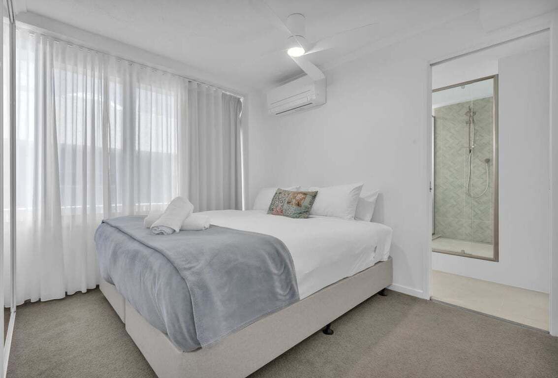 A bedroom with a bed, a window with curtains, an air conditioning unit, and a shower area.