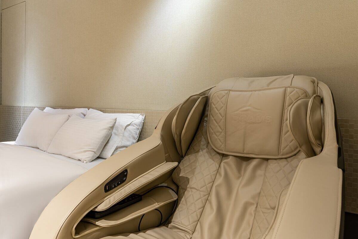 A massage chair in a hotel room with a bed and a tiled wall.