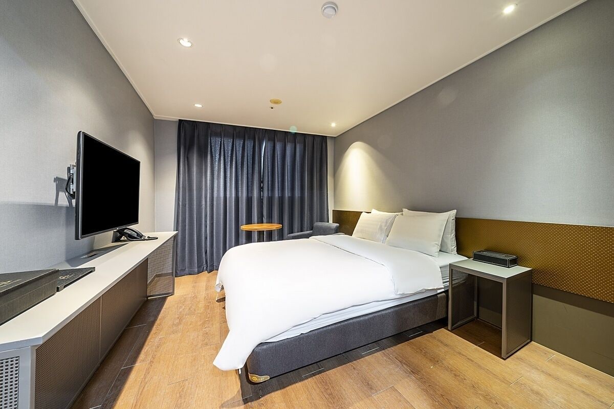 A modern hotel room with a bed, bedside table, TV, and a desk with a computer.
