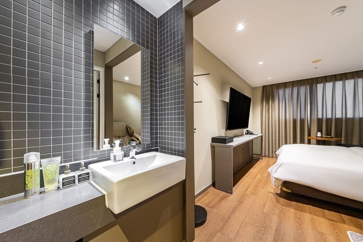 A hotel room with a large mirror, a sink, a television, a bed, and a window with curtains.