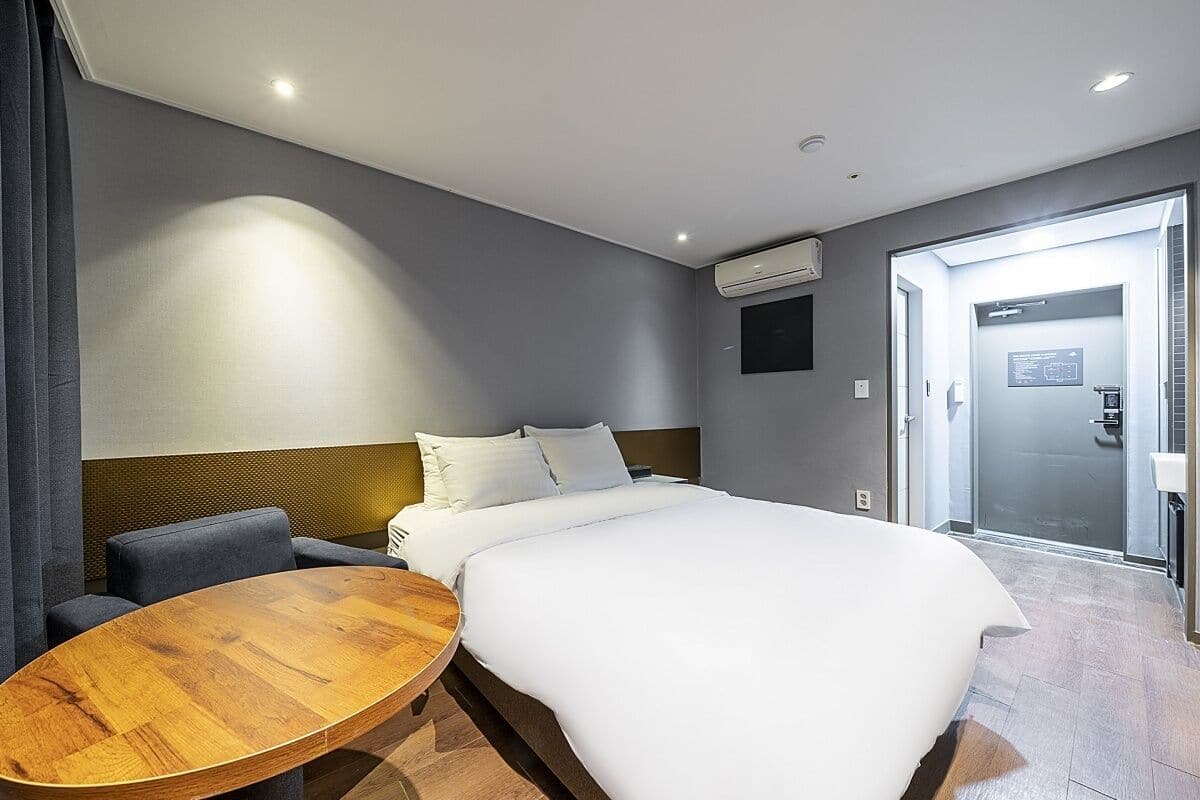 A modern hotel room with a large bed, a round wooden table, a sofa, and a glass door leading to a bathroom.