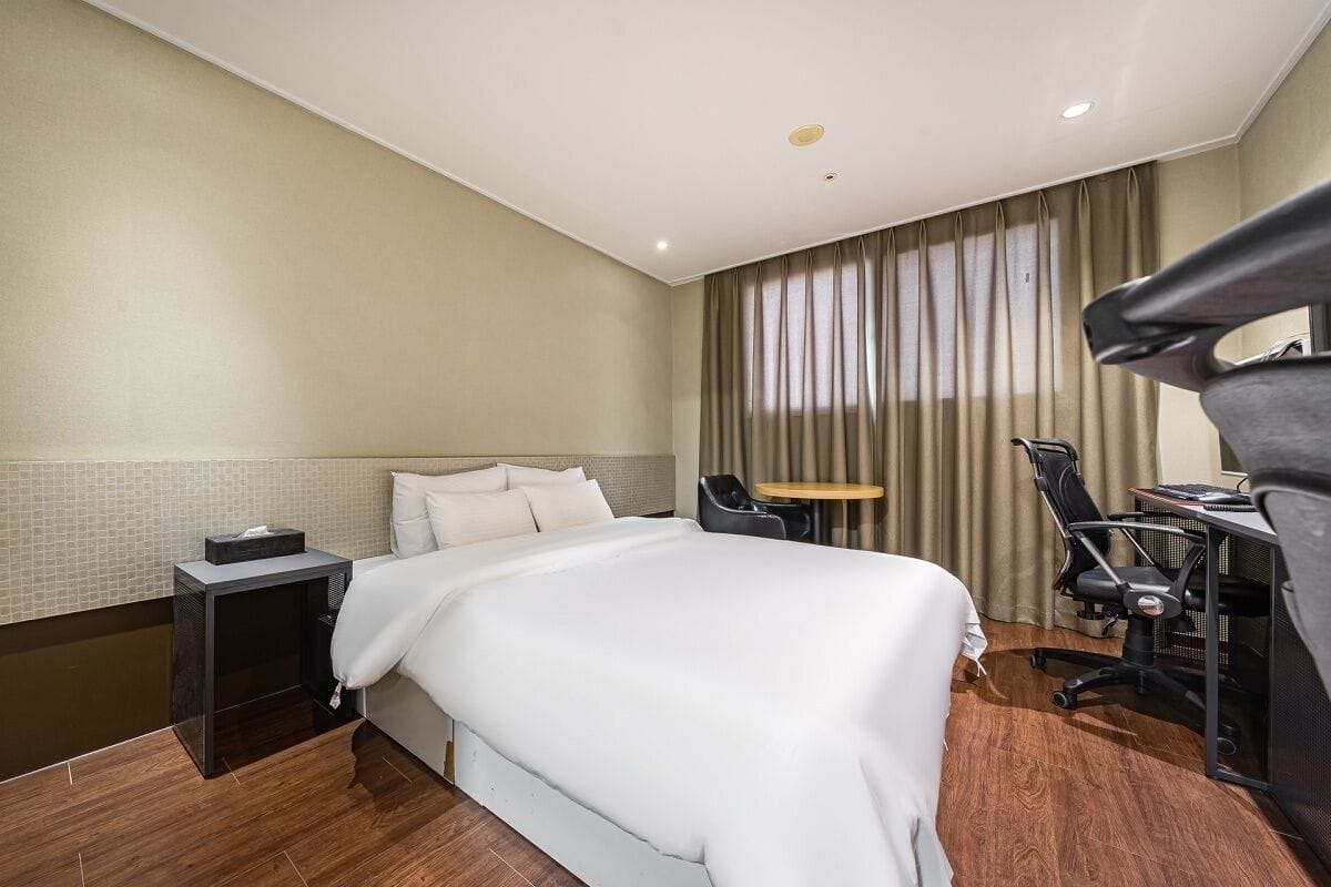 A hotel room with a large bed, a desk with a chair, a small table, and a curtain-covered window.