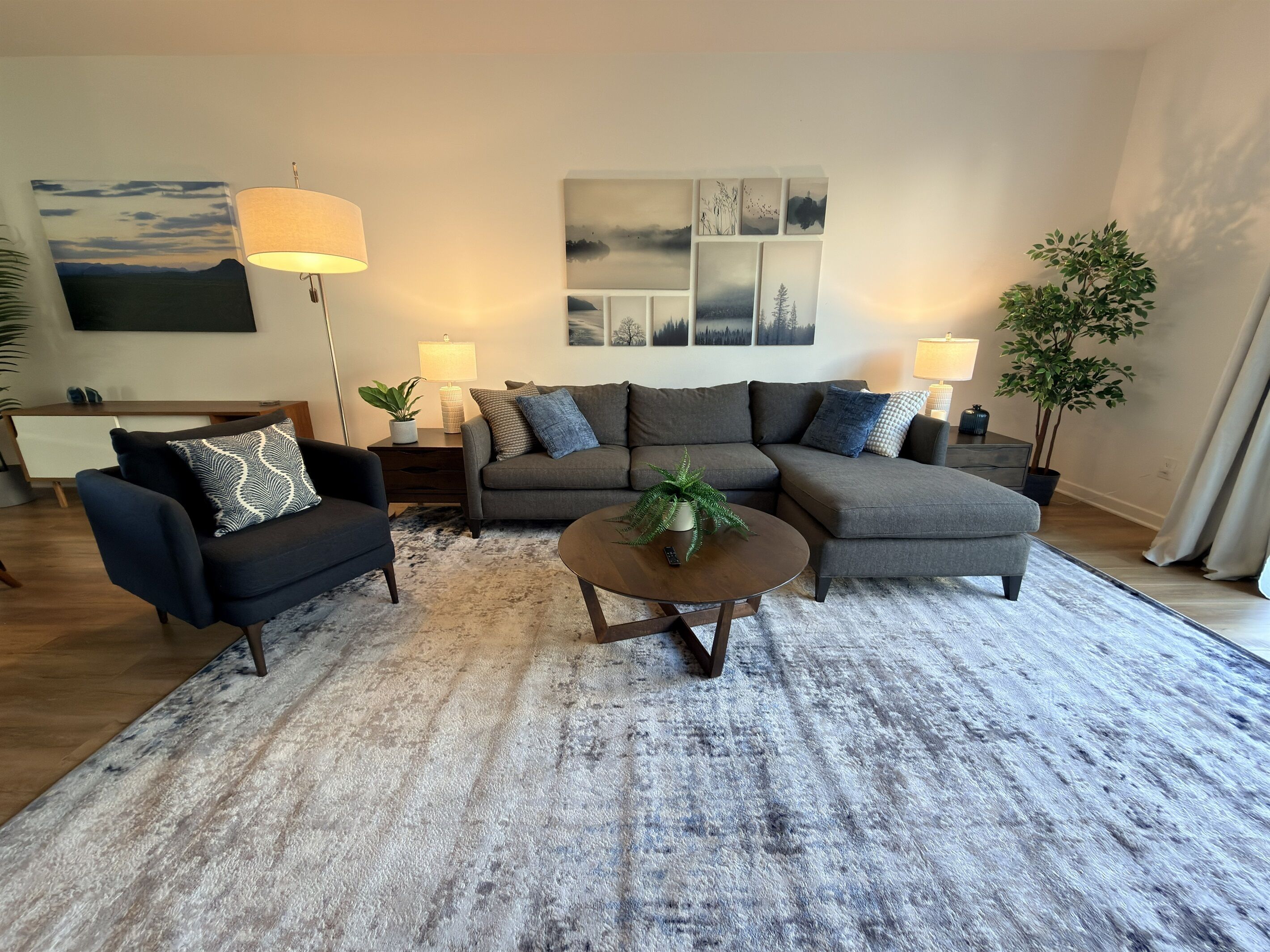 A living room with a sectional sofa, a round coffee table, and a small side table. There is a flat-screen TV mounted on the wall, a potted plant, and a large window with curtains.
