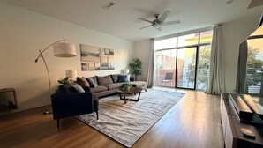 En rummelig stue med stor sofa, sofabord og gulvlampe. Loftsventilator og stort vindue giver naturligt lys.