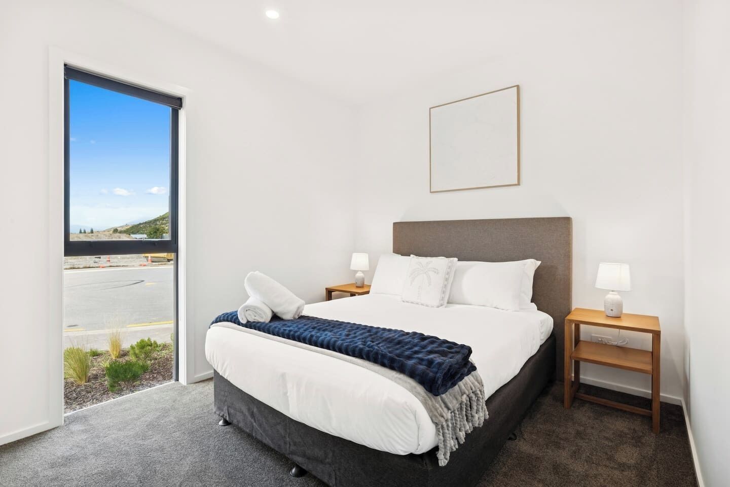 A modern hotel room with a large bed, two bedside tables, a view through a sliding door, and a framed picture on the wall.