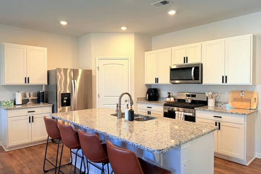 Cocina moderna con armarios blancos, encimeras de granito, electrodomésticos de acero inoxidable y una isla central con taburetes de bar.