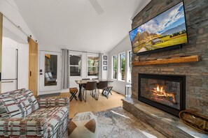 A modern living room with a fireplace, a television mounted on a stone wall, and a dining area with a table and chairs. - Moondrift Retreat - pet friendly, hot tub, community amenities, ranch style home (Newland)