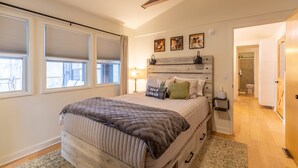 Quarto com uma cama grande, uma mesa de cabeceira com abajur e cabeceira decorada com arte. Há uma janela com persianas e uma porta que dá para o banheiro.