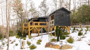 Uma cabana com deck de madeira e fachada escura, cercada por árvores e neve.