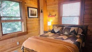 Quarto de cabana de madeira com cama, mesa de cabeceira, abajur e um quadro na parede.