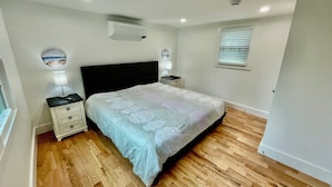 A bedroom with a bed, bedside tables, a wall-mounted air conditioner, a window with blinds, and a circular wall decoration.