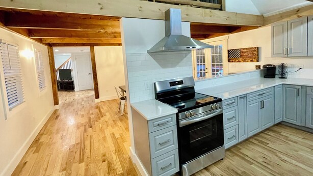 A modern kitchen with a stainless steel stove, a large oven, and light grey cabinets. There is a wooden ceiling and a staircase leading to an upper level.