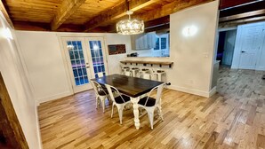 A dining area with a wooden table and white chairs, a kitchen in the background, and a wooden ceiling.
