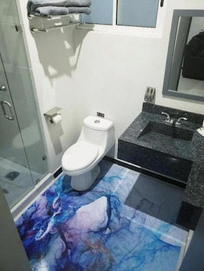 A bathroom with a white toilet, a glass shower enclosure, a black sink with a marble countertop, and a colorful abstract floor design.