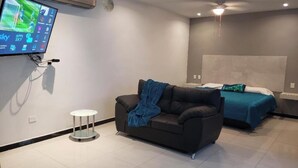 A modern hotel room with a flat-screen TV mounted on the wall, a sofa, a bed with a blue bedspread, and a small round table. - PRIVATTO SUITES (Saltillo)