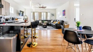Eine moderne Küche mit einer zentralen Kochinsel, ein Essbereich mit rundem Tisch und Stühlen, ein Wohnzimmer mit Sofa und Couchtisch sowie ein Deckenventilator.