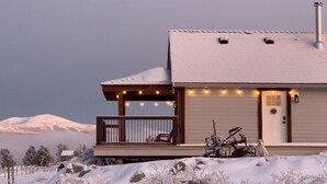 A cabin with a snow-covered roof, a wooden deck with a railing, and a bicycle parked outside. - Come to the Mountains, Stay on the Farm - Cozy Farm-Stay Cottage (Bridesville)