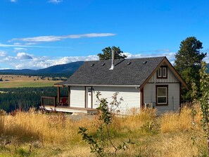 A single-story house with a gabled roof, a chimney, and a porch with a red chair. The house is surrounded by tall grass and trees, with a view of a mountain range in the background.