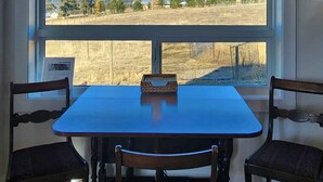 A dining area with a table and four chairs, a window with a view of a field and mountains, and a framed picture on the table. - Come to the Mountains, Stay on the Farm - Cozy Farm-Stay Cottage (Bridesville)