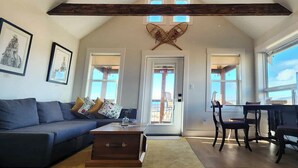 A spacious living room with a high ceiling, a dark wooden beam, a large window, a sofa, a coffee table, and a dining area with chairs. - Come to the Mountains, Stay on the Farm - Cozy Farm-Stay Cottage (Bridesville)