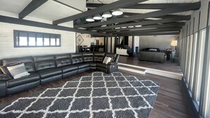 Un amplio espacio de sala con un sofá seccional de cuero negro, un tapete estampado y una cocina al fondo.