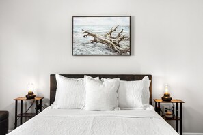 Ein Schlafzimmer mit einem großen Bett, zwei Nachttischen mit Lampen, einem gerahmten Bild von einem Baum an der Wand und einer weißen, strukturierten Bettdecke.