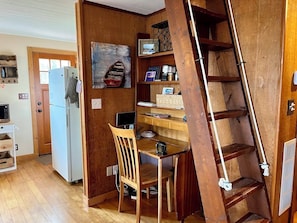 A compact kitchen with wooden cabinetry, a white refrigerator, a wooden dining table with a chair, and a wooden ladder.