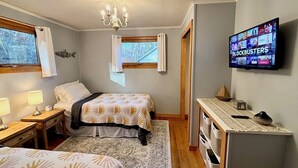 A bedroom with two beds, a TV displaying 'BLOCKBUSTERS', a wooden dresser, and a window with curtains. - Kayak on Prospect Lake, Paddle Boards, Swim Dock+! (Egremont)