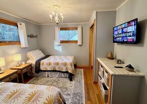 A bedroom with two beds, a TV displaying 'BLOCKBUSTERS', a wooden dresser, and a window with curtains.