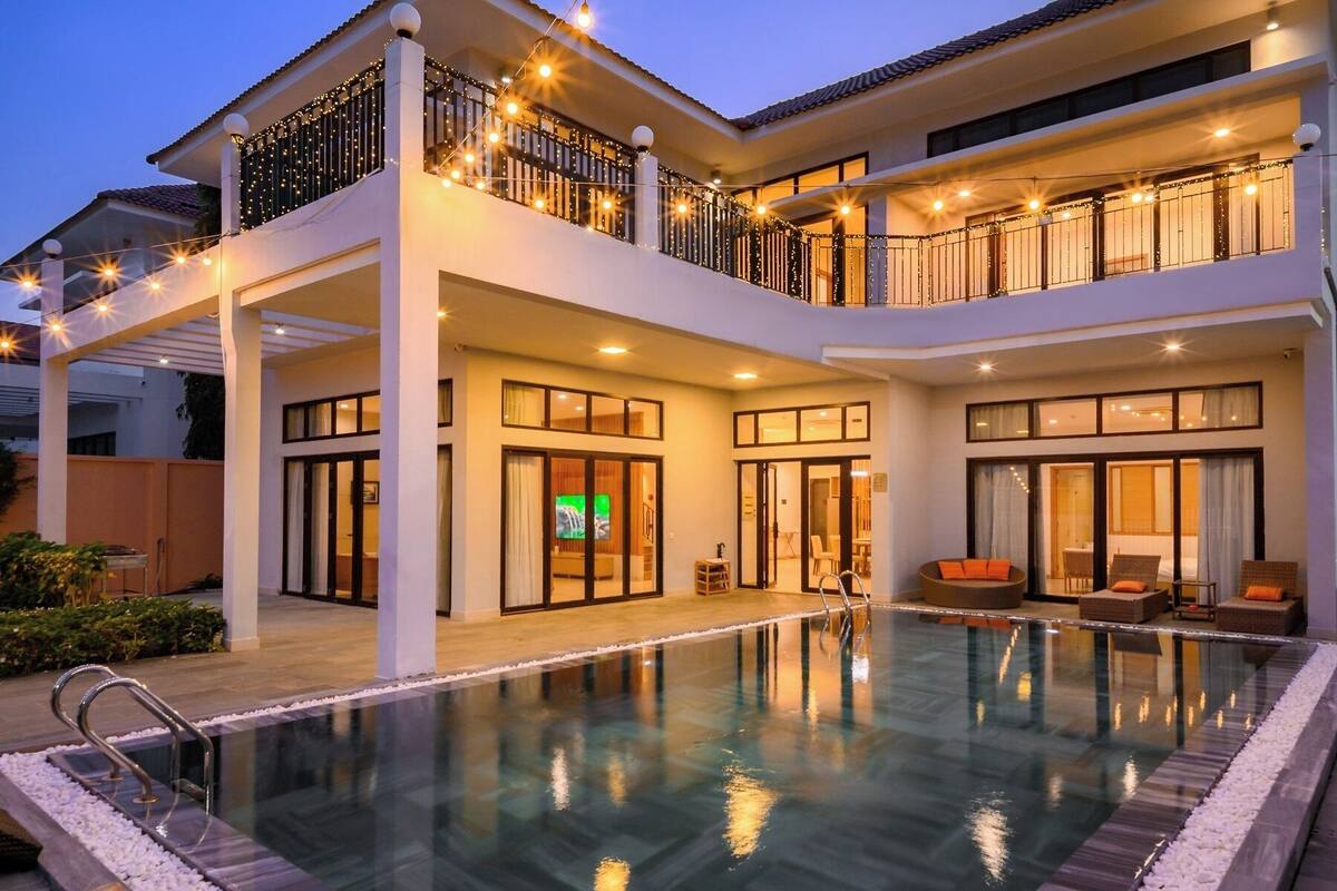 A two-story building with a swimming pool, balconies with string lights, and a visible entrance with glass doors.
