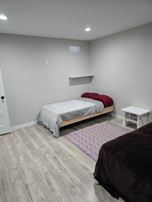 A bedroom with a bed, a small white nightstand, a patterned rug, and a shelf above the bed.