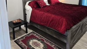 Un dormitorio con una cama de madera, una mesita de noche pequeña, un tapete rojo y una lámpara en el techo.