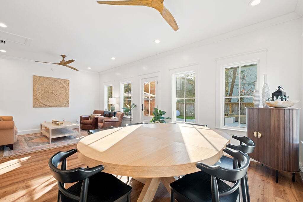 A spacious room with a large round wooden table surrounded by chairs, a ceiling fan, and large windows allowing natural light.