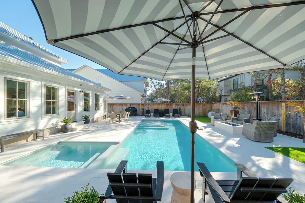 Un patio trasero con una piscina rectangular, sillas de descanso y un paraguas grande.