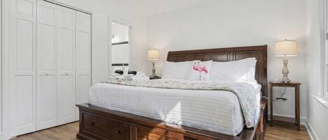 A bedroom with a wooden bed, white bedding, a nightstand with a lamp, a dresser, and a closet with white doors.