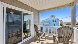 A covered porch with two wooden chairs, a sliding glass door, and a view of a blue house in the background. - 8388 Family Island Retreat | Steps to Beach (Avon)