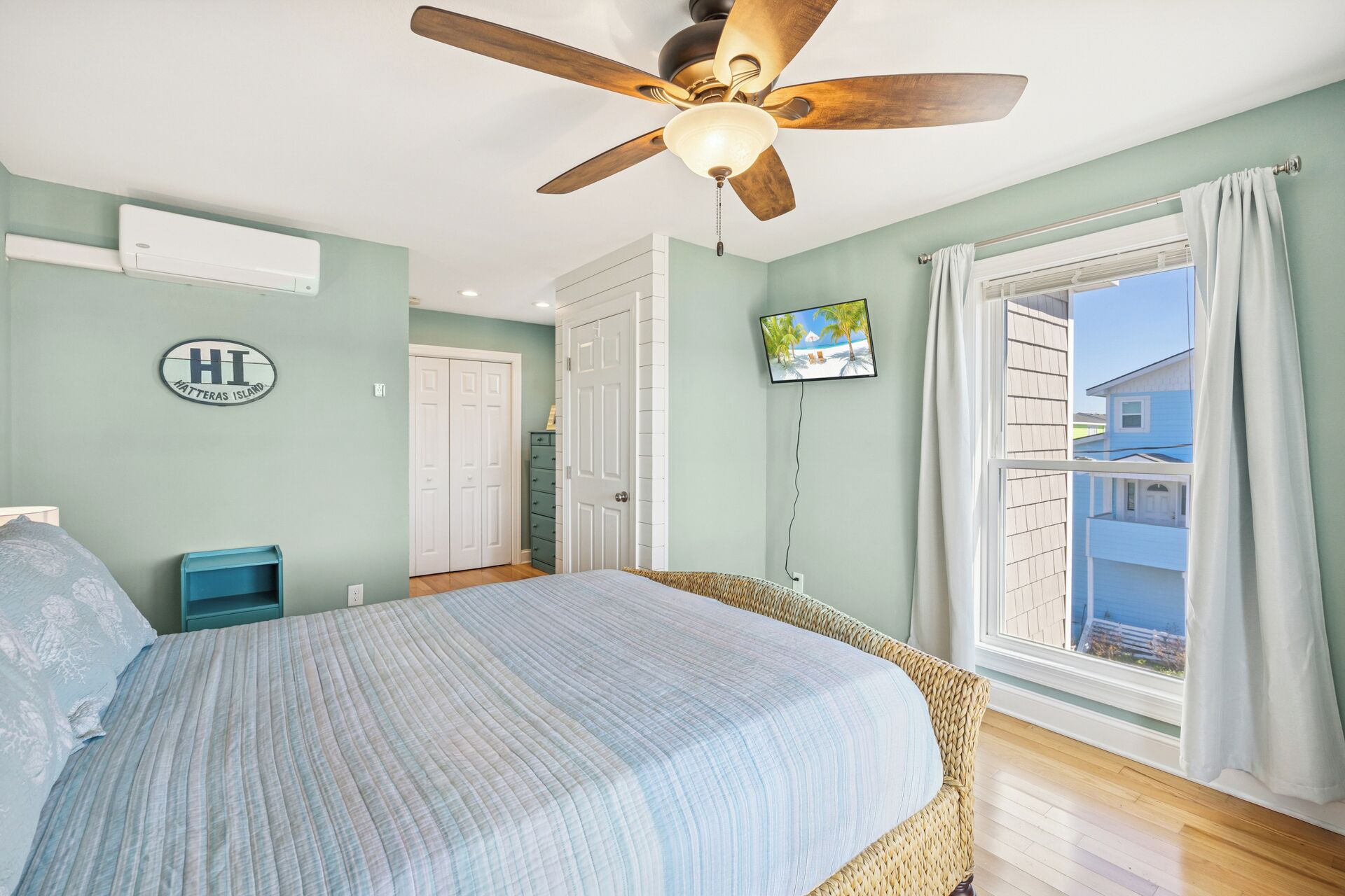 A bedroom with a bed, a ceiling fan, a TV, and a view of a house through the window.