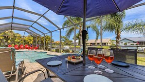 A covered outdoor area with a pool, lounge chairs, and a table set for dining. - Canalfront Island Escape with Pool, Lanai & Dock - Antigua Villa - Roelens (Cape Coral)
