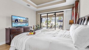 A hotel room with a bed, a TV, a desk with a bottle and glasses, and a view of the outside. - Canalfront Island Escape with Pool, Lanai & Dock - Antigua Villa - Roelens (Cape Coral)