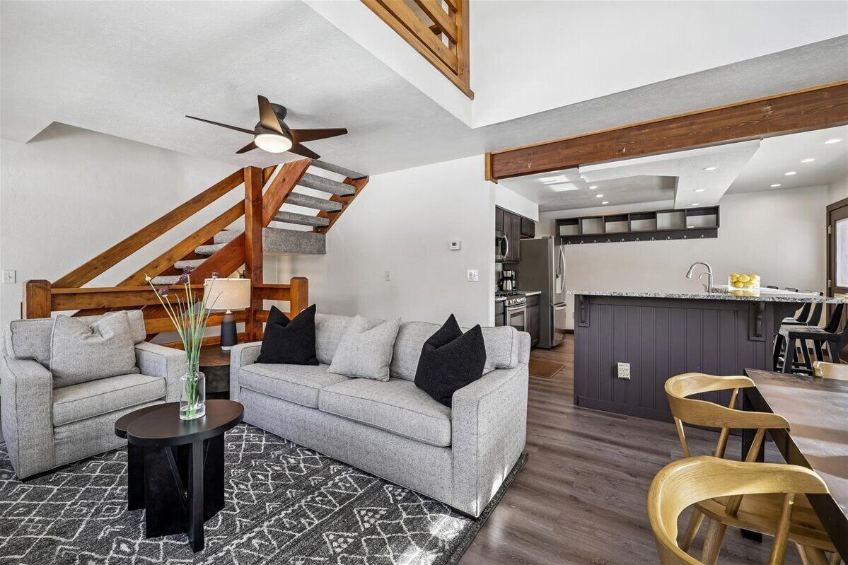 A modern living room with a grey sofa, black cushions, a small black side table, and a patterned rug. A wooden staircase with a ceiling fan leads to a kitchen area with a bar counter and wooden chairs.