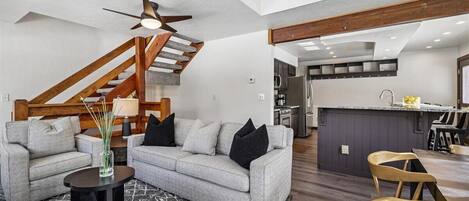 A modern living room with a grey sofa, black cushions, a small black side table, and a patterned rug. A wooden staircase with a ceiling fan leads to a kitchen area with a bar counter and wooden chairs.