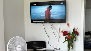 Una sala de estar moderna con un televisor de pantalla plana montado en la pared, una consola de madera con un jarrón de flores, un ventilador de pie y un sofá gris.